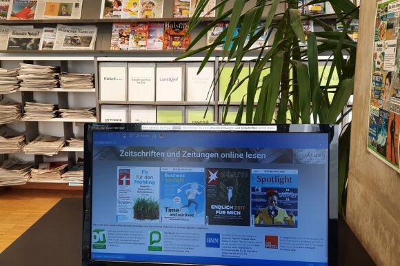 Vorne ein Bildschirm mit Online-Zeitschriften, dahinter ein Regal mit analogen Zeitschriften.