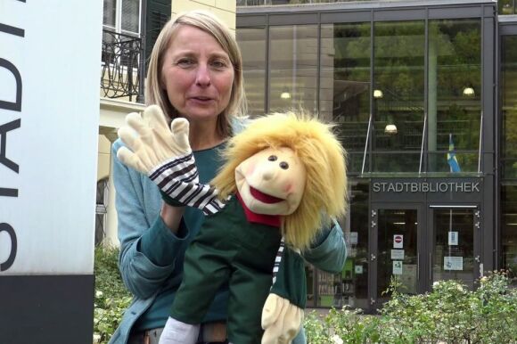 Frau mit Stoffpuppe winkt vor Stadtbibliothekgebäude