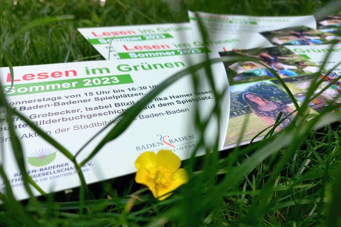 Meherere Schilder zum "Lesen im Grünen" auf Wiese ausgebreitet, davor gelbe Butterblume.