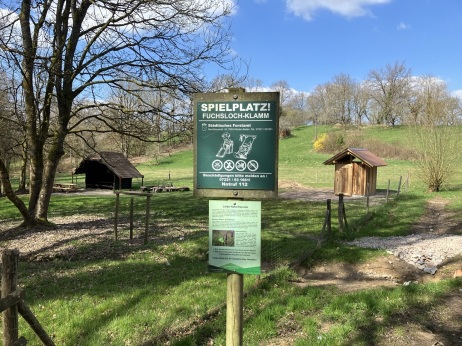 Ein Schild mit der Aufschrift "Spielplatz" und im Hintergrund der Grillplatz Fuchsloch. 