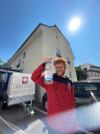 Ein Mann mit rotem Kapuzenpullover steht in der Sonne und hält eine Wasserflasche in die Luft.