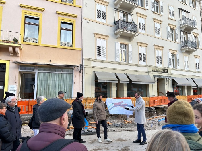 Erster Bürgermeister Alexander Wieland und Projektleiter Vincenz Wienk-Borgert halten einen Plan in den Händen und stehen mit weiteren Personen auf der Baustelle am Hindenburgplatz. 