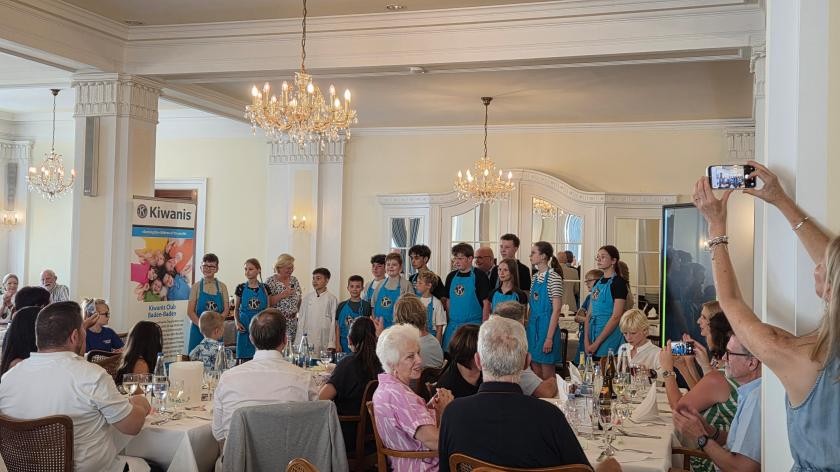 Eine Gruppe junger Menschen in blauen Kochschürzen steht vor einem Publikum, dass an gedeckten Tischen sitzt.